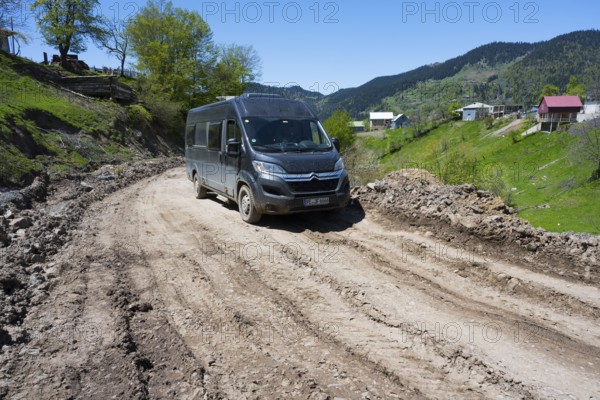 A van drives down a muddy road surrounded by green hills and individual houses, campers on the way