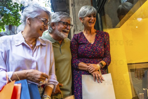 Senior friends laughing together while window shopping on a city street, carrying multiple shopping
