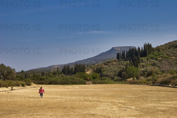 A child runs across a dry meadow to distant mountains, under a clear sky with cypresses in the