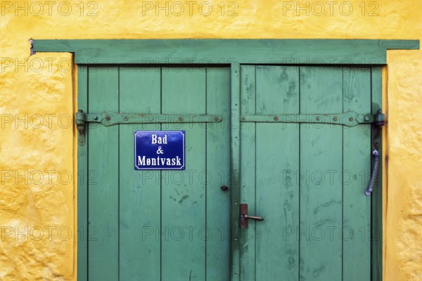 Green wooden door in a yellow wall, blue sign with inscription in Danish language, bathroom and