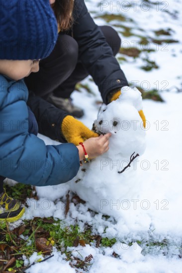 Young boy and mother collaborating to build a small snowman, carefully adding details like eyes and