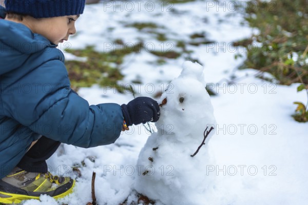 Young boy playing in the snow, carefully placing a small natural element on a snowman he is
