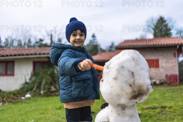 Smiling little boy in warm winter clothes builds a snowman in the yard, holding a carrot to place