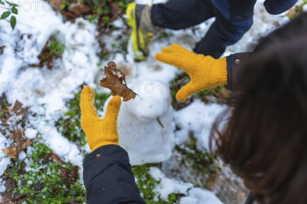 Child wearing yellow wool gloves decorates a small snowman with a brown leaf in a snowy forest,