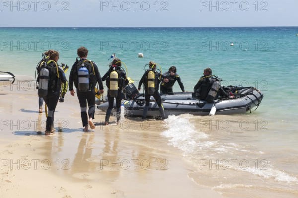Recreational divers walk across beach to inflatable boat in diving suits with scuba tanks on their