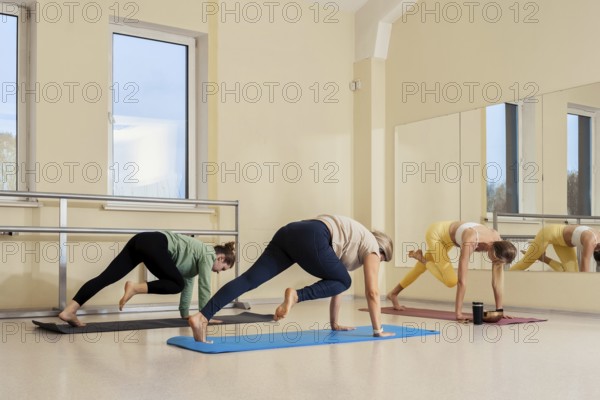 Three women engage in a fitness session in a sunny studio. They do yoga poses on colorful mats with