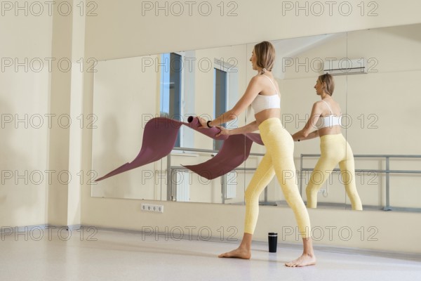 A woman prepares for a yoga session in a bright studio, unfolding her mat on the smooth floor. She