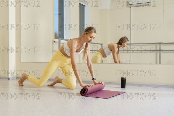 A woman in comfortable activewear rolls out her yoga mat in a well-lit studio. She is focused and