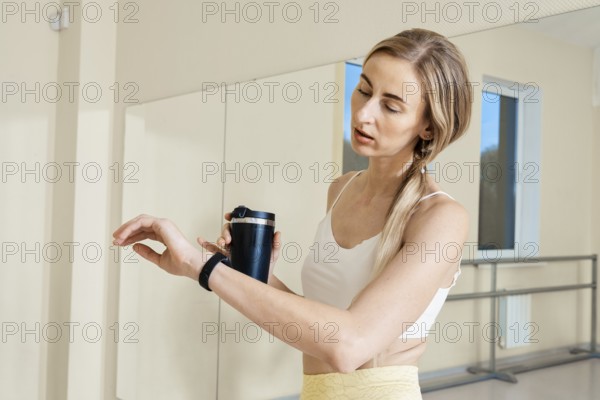 A woman in a fitness studio checks her smartwatch while holding a black drink container. She wears