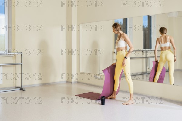 In a well-lit studio with large mirrors, a woman stands barefoot holding a yoga mat. She is dressed