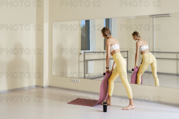 A woman in fitted yoga attire is standing in a yoga studio, preparing to practice. She stretches