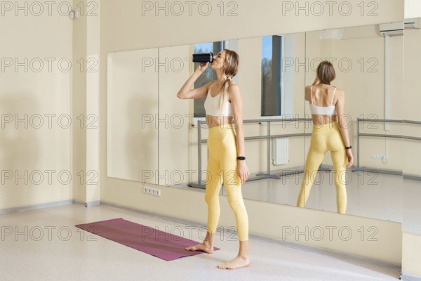 In a bright yoga studio, a woman in yellow leggings and a sports bra takes a sip from a water