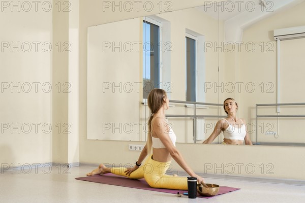 A woman practices yoga in a well-lit studio. She sits in a split position on a yoga mat while