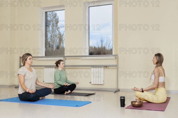 Three women are seated on yoga mats in a bright studio. They are focusing on meditation, with one