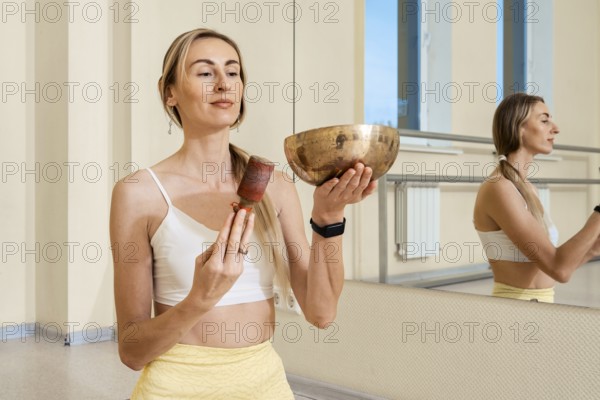 A woman stands in an airy studio, holding a singing bowl in one hand and a mallet in the other. She