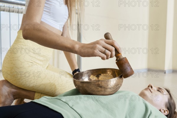 In a calm wellness studio, a sound healer plays a singing bowl on a person lying calmly on a mat.