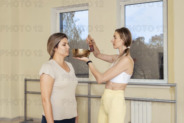 In a serene studio, a woman stands calmly while another person gently uses a singing bowl above her