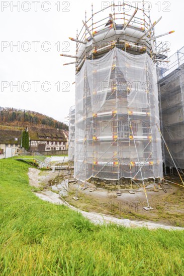 Large tower in scaffolding with surrounding meadow, trees and buildings in the background,