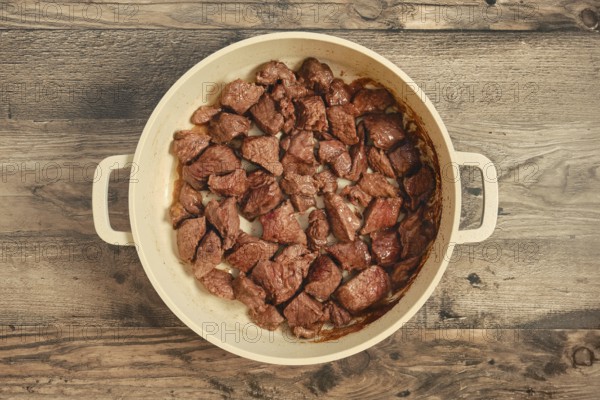 Chunks of beef are searing in a large pot on a weathered wooden table. The meat is cooked to a rich