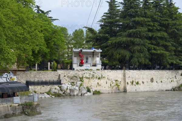 A riverbank cable car station surrounded by lush trees and adjacent buildings, cable car cabin,