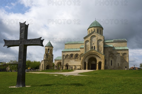 Historic church with cross in the foreground, cloudy sky surrounded by green landscape, Bagrati
