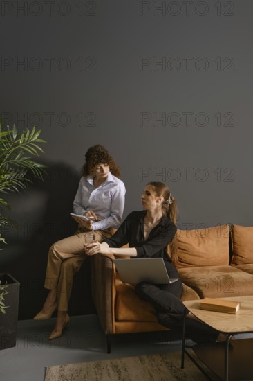 In a stylish living room, two women engage in a collaborative work meeting. One writes notes while