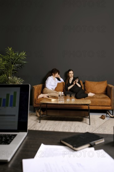 Two individuals are seated comfortably on a stylish couch, one looking at a phone while the other