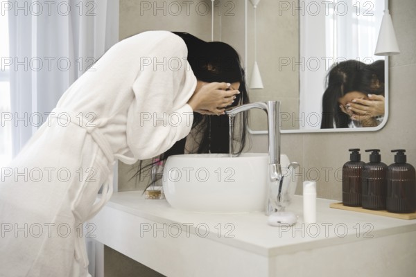 A woman in a white bathrobe leans over a stylish sink in a contemporary bathroom, splashing water