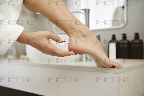 A person gently massages their foot while resting on a sink in a stylish bathroom. Soft morning