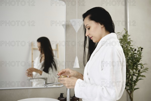 A woman with long dark hair stands at a sleek sink in a contemporary bathroom and opens a jar of