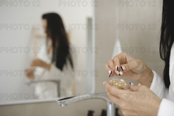 In a stylish bathroom, a woman in a robe is holding a small jar with skin vitamins and preparing to