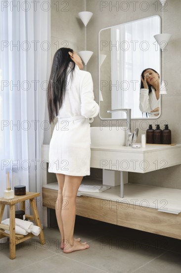 Barefoot woman wearing a white robe stands by a bathroom sink, facing the mirror. She appears to be
