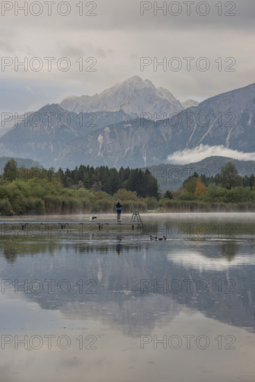 Woman (40-50) with dog on a wooden footbridge at sunrise, Hopfensee, near Füssen, Ostallgäu, behind