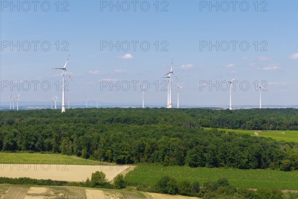 Several wind turbines on the horizon above a forest and fields, under a blue sky, near Volkach,