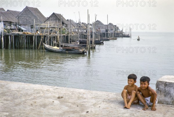 Two boys sitting on quayside next to attap houses of fishing village on stilts, Singapore,