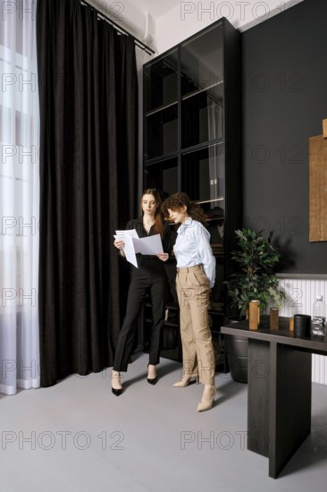 Two professional women stand in a sleek office, reviewing important documents. Sunlight filters
