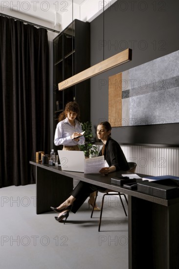 Two businesswomen are engaged in a discussion at a sleek office desk. One is seated while reviewing