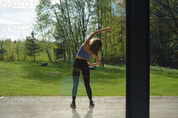 Rear view at a woman tilting her body to the sides on a wooden deck, surrounded by lush greenery