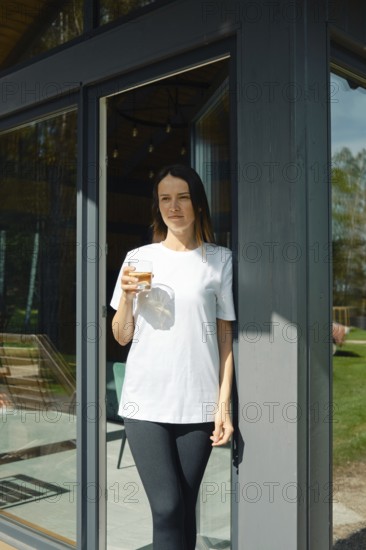 A young woman stands leisurely at the entrance of a stylish house, holding a drink in her hand.