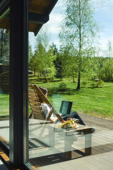 A person enjoys a peaceful moment outdoors, seated in a wooden lounge chair. Surrounded by lush