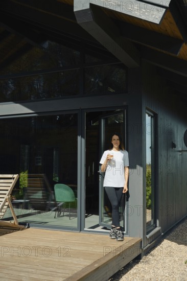 A woman stands on the wooden deck of a stylish cabin, holding a drink while enjoying the serene