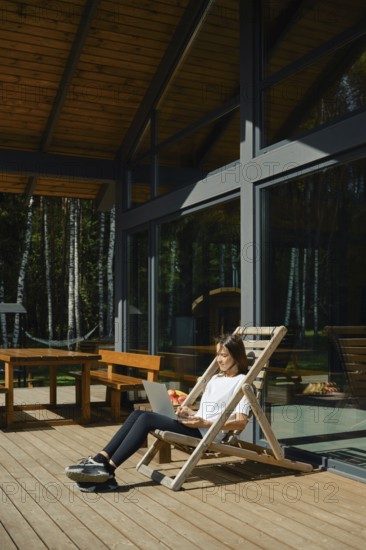A woman sits on a wooden chair outdoors and typing on a laptop. She enjoys her vacation in modern