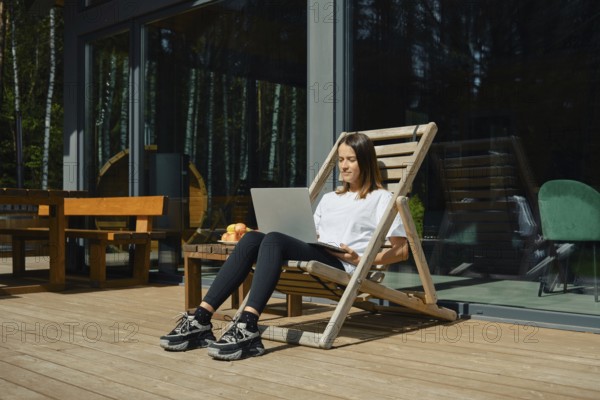 A woman is sitting in a wooden lounge chair, engaged with her laptop on a sunny deck. She enjoys a
