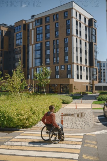 A man using a wheelchair navigates a crosswalk in a modern urban setting. The sun shines brightly,