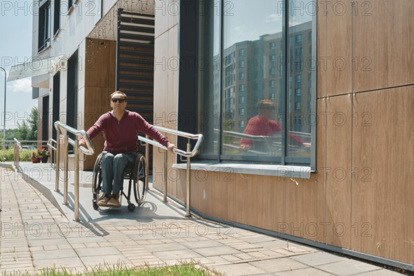 Individual in a wheelchair is utilizing a ramp to exit a modern building with large windows. The