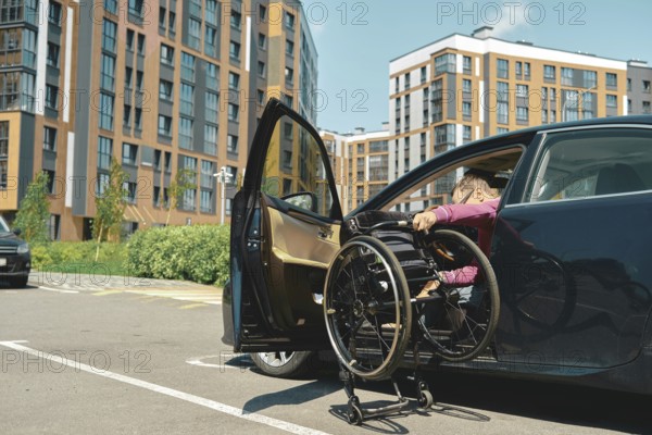 A person is maneuvering a wheelchair into a car parked in a contemporary urban area. A man unfolds