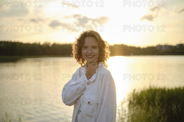 A woman stands by the lake, smiling as the sun sets behind her, casting a warm golden glow on the