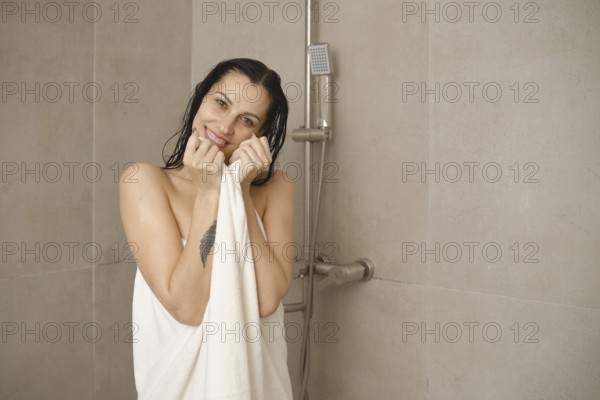 A woman stands in a modern bathroom, smiling while holding a towel close to her body. The warm