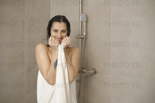 A woman with wet hair smiles while holding a towel in a modern bathroom. The tiles are light, and a