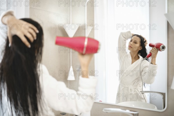 A woman is using a hair dryer to style her wet hair while standing in front of a large mirror. The